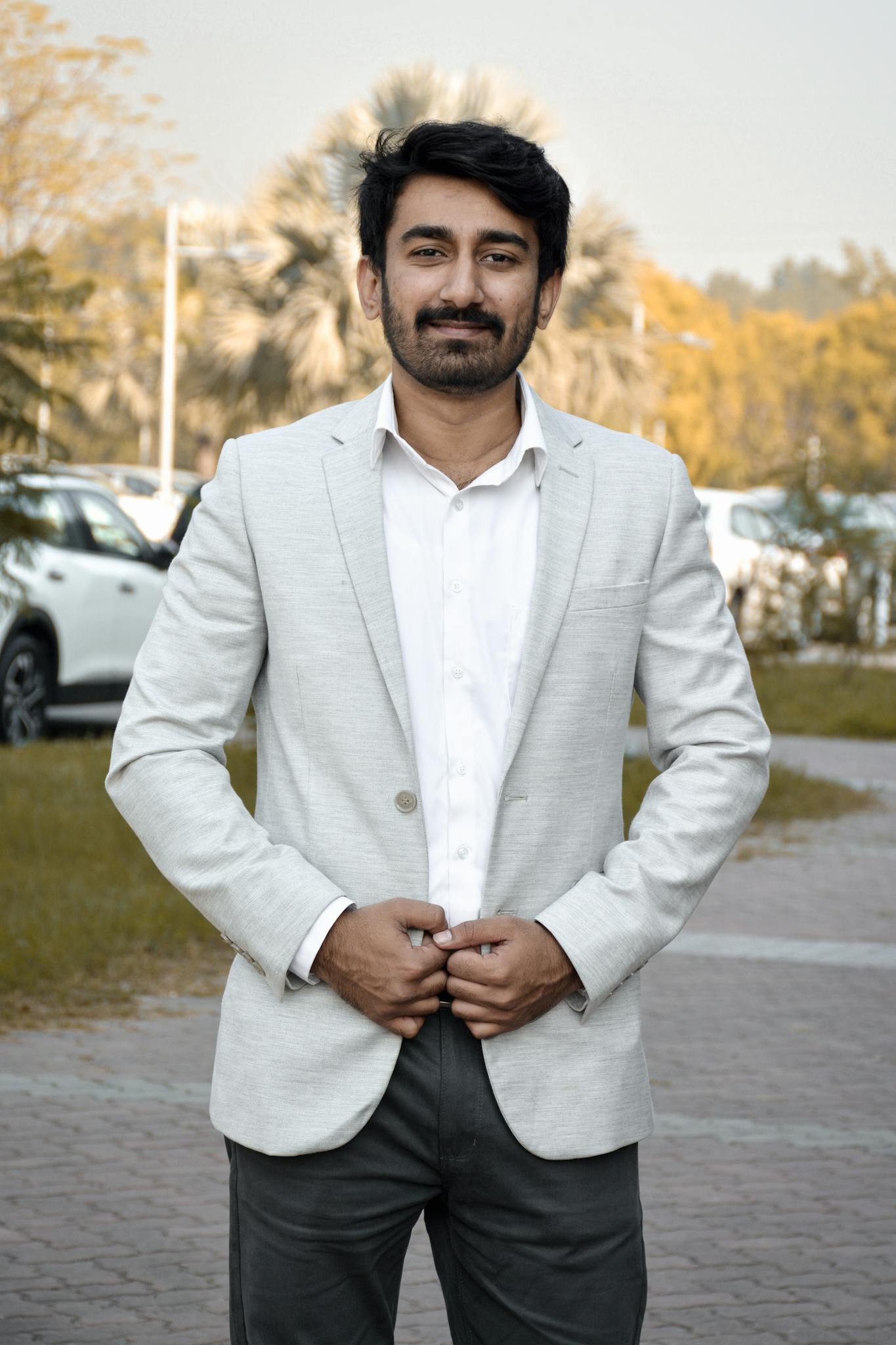 Confident man in white suit standing outdoors with cars and trees in the background.