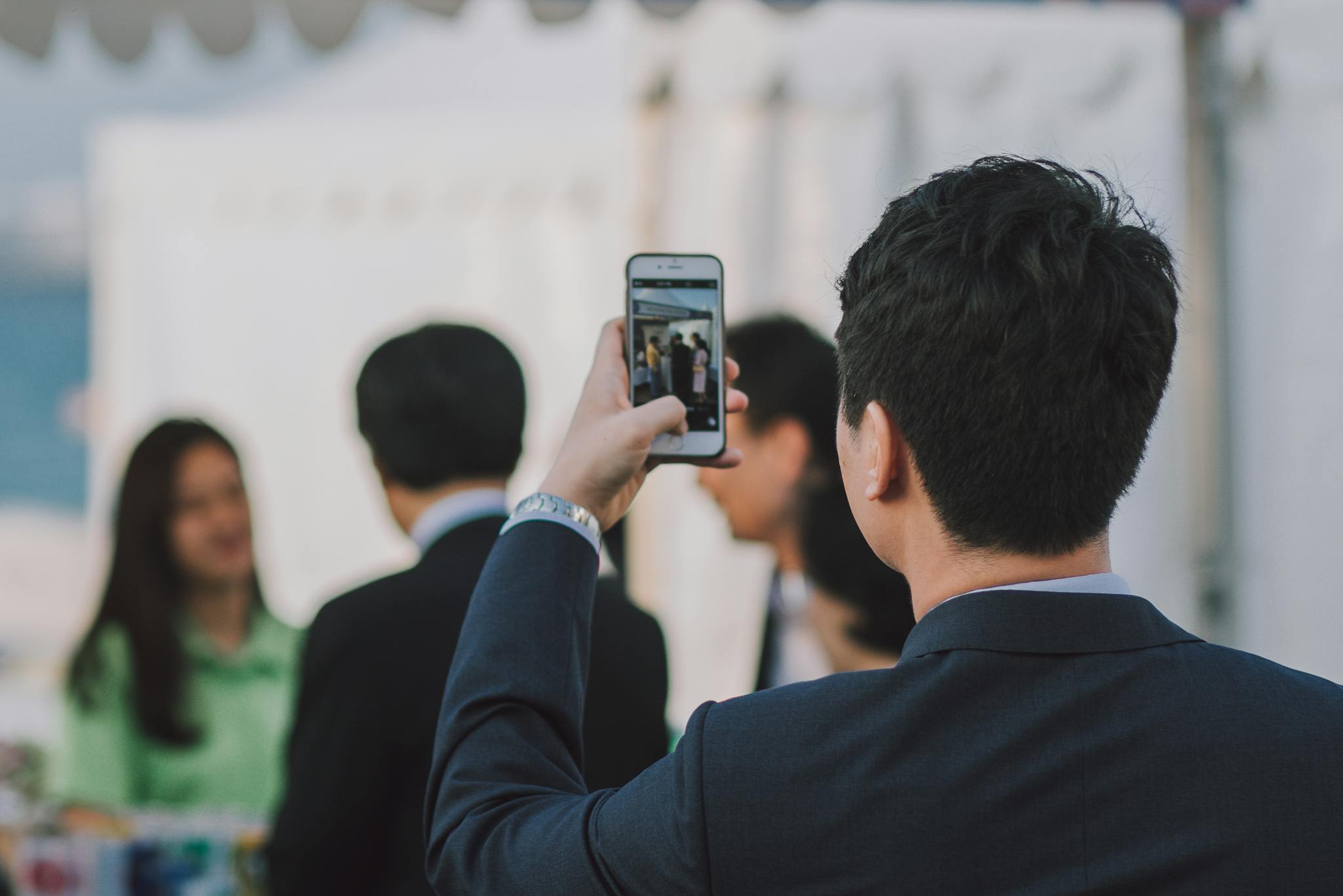 Man in suit photographing colleagues with smartphone during business event outdoors.